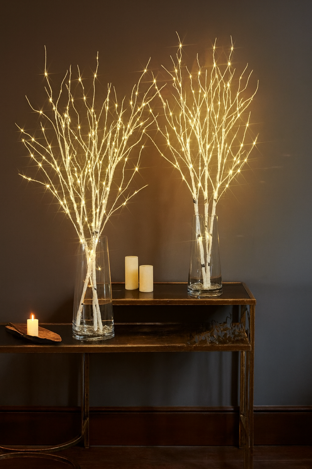 Two illuminated twig decorations in glass vases on a wooden table against a dark wall.